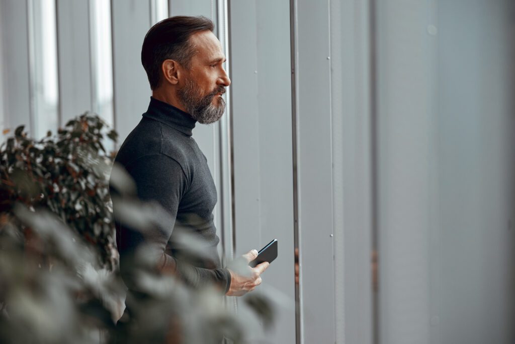 It feels good when you become an AI Productivity Hero. Image description: Bearded man with mobile phone looking out the window in the room